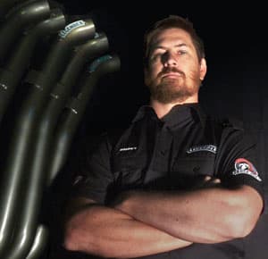 Barney Lewis in a Legendex workshop shirt, arms folded, beside a set of exhaust headers